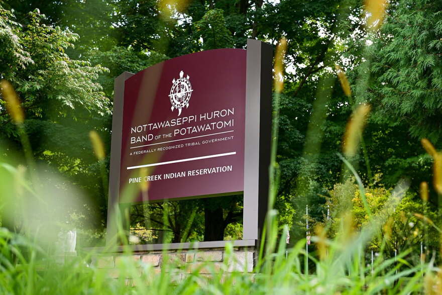 A dark red sign for the Nottawaseppi Huron Band of Potawatomi's Pine Creek Reservation can be seen in a shot that's looking up from the ground through grass, with trees in the background behind the sign