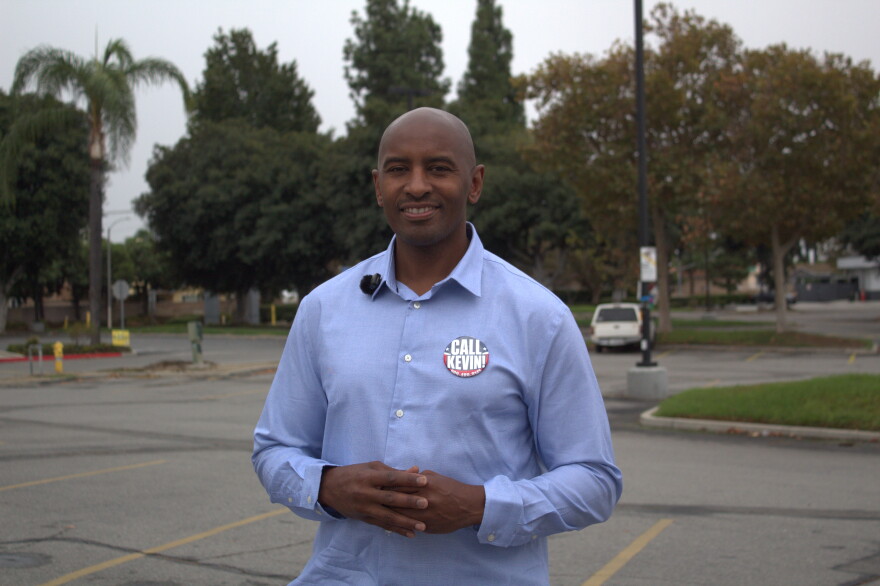 A man with a tennis-ball-sized-pin on his shirt that reads "CALL KEVIN!," stands in a parking lot with his hands together at his torso