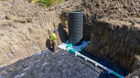 A man in a bright yellow shirt stands near a tank and a long pipe, surrounded by a pile of dirt.