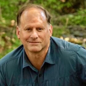 Todd Davis, with a slight smile, outdoors in a wooded area, wearing a blue shirt