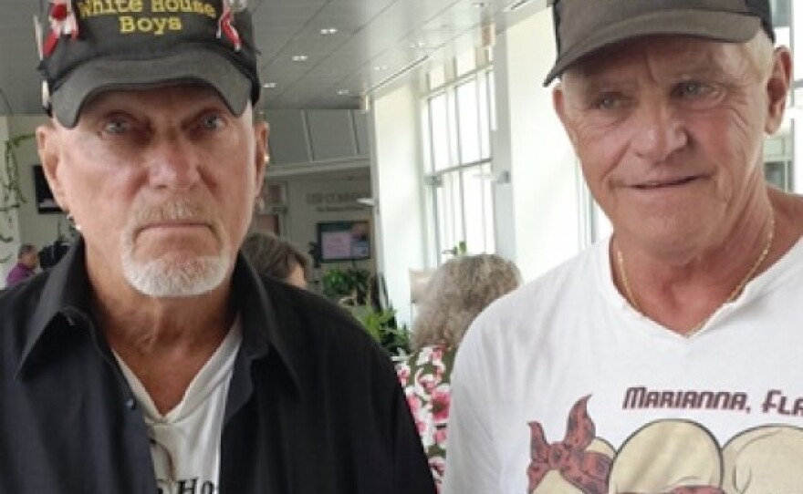 Two older men, wearing baseball caps that read "The White House Boys," hold marbles in their hands. 