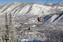 Skiers and snowboarders ride up a ski lift with snowy hills and a town in the background.