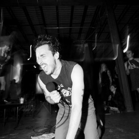 Sal from Gatto Black on his knees, singing passionately into a microphone in a black and white photo.