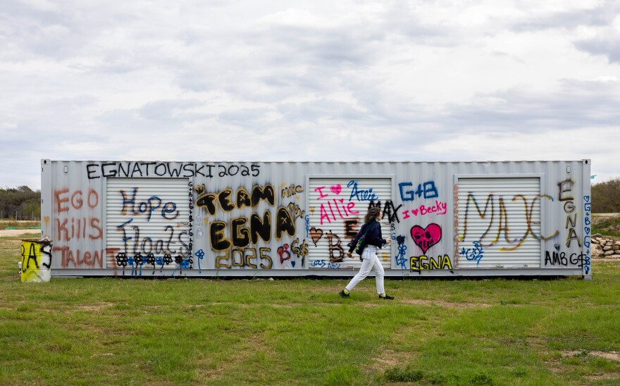 A shipping container spray painted with graffiti sits in a field as a person walks past.