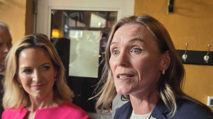In the foreground, Winnie Brinks wears a navy blazer, American flag pin, and gold necklace. Jocelyn Benson stands beside her in a bright pink dress, smiling and looking toward Brinks. The two women are in a warmly lit room.
