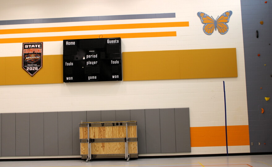 A gymnasium wall featuring a digital scoreboard, a championship banner, a butterfly wall decal, and a portion of a climbing wall.