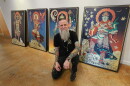 Man with tattoos all over his arms and a long grey beard squats in front of his artwork 
