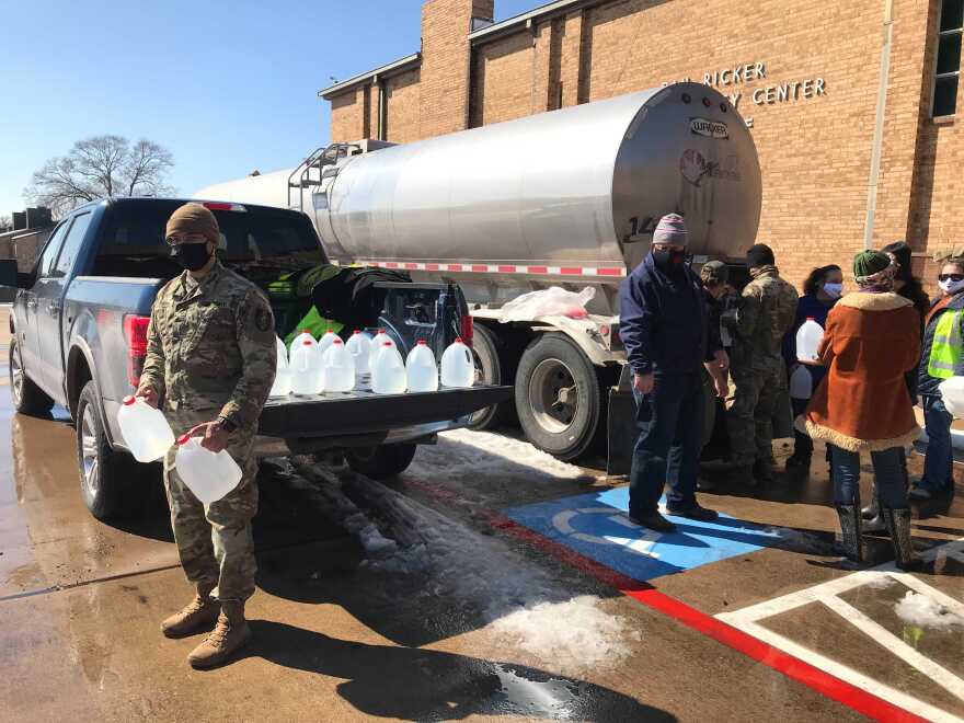 A man in army fatigues holds jugs of plastic water in front of a pickup truck filled with water jugs and next to a tanker truck, where several people are lined up to fill up jugs.