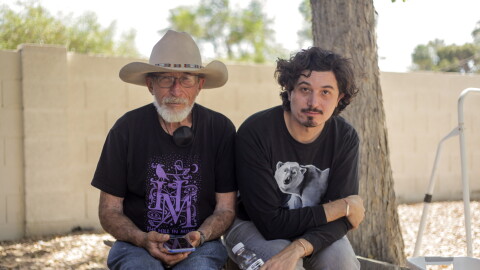 An elderly father and his adult son sit next to a tree in a backyard surrounded by a block wall. 