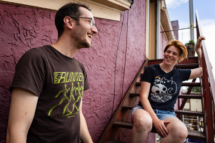 Photo of two people on metal stairs leading up the side of a red house to a front door. One person is standing while the other is sitting on the stairs. Both are smiling brightly.