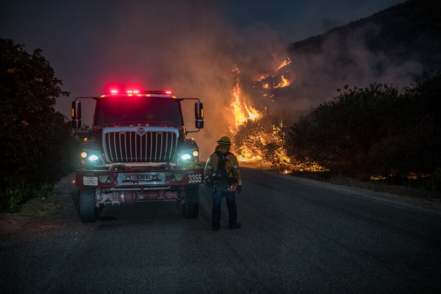 A CAL FIRE firefighter stands next to a fire engine as the Fairview Fire burns close by.
