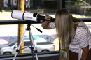 A woman's blonde straight hair drapes down, as she bends to peer through a white telescope, grasping it with her left hand. The instrument, aimed out a large glass window, has black lettering that reads Hexeum.