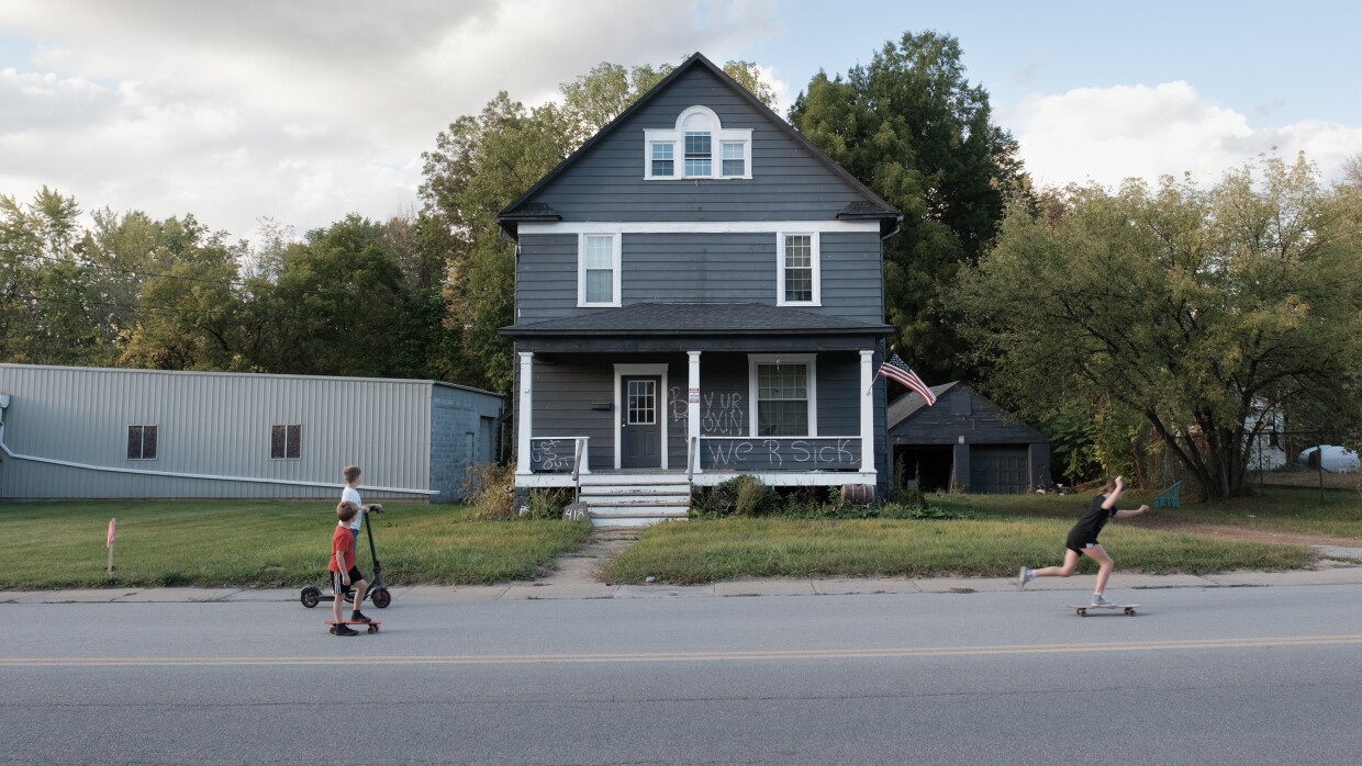 A gray house with the words "We R sick" written on the front as children on skateboards and a scooter roll by on the street.