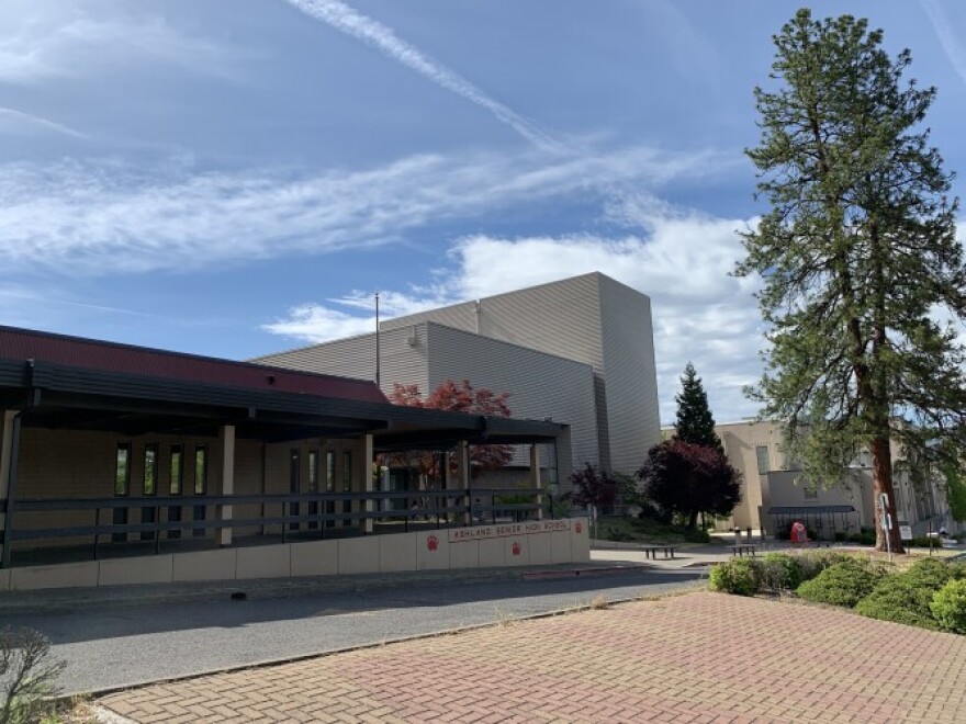 A large gray building with trees and bricks in front of it. Behind it is blue sky. 
