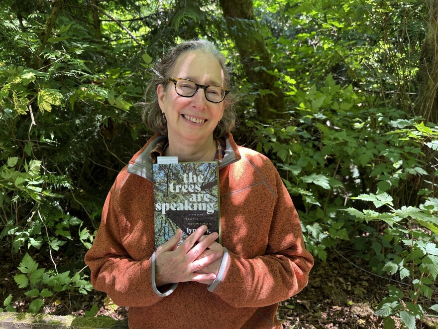 Author Lynda V. Mapes stands in the forest at Seattle's Carkeek Park, smiling. She is holding a copy of her new book, The Trees are Speaking.