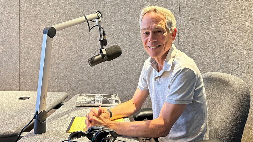 A man sits with his hands folded on a table in front of a microphone on an arm inside a professional radio studio.