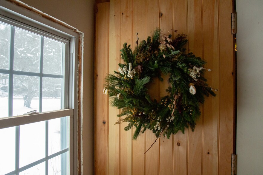 A wreath hangs on a wood-paneled wall, next to a window. Outside, daylight filters in and there is snow on the ground.