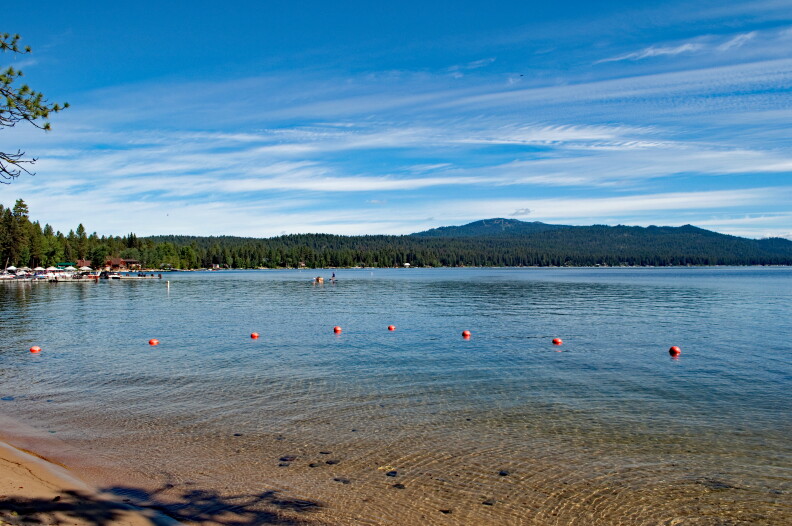 A blue lake with some red buoys near the shoreline. You can see trees and mountains in the background of behind the water.