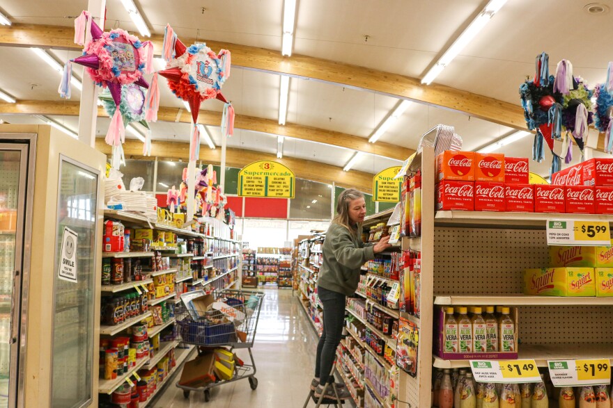 A woman stands on a step stool to place grocery items on a shelf.