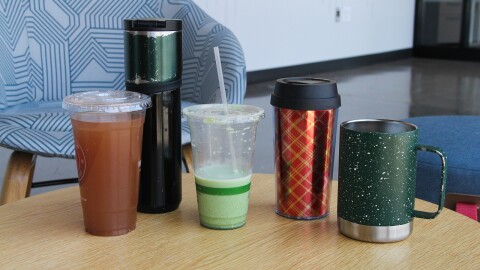 Five coffee cups sit on a table: a clear plastic single-use cup, a tall reusable to-go cup, a half-empty, clear plastic single-use cup, a smaller reusable to-go mug, and a metal mug with a handle. 