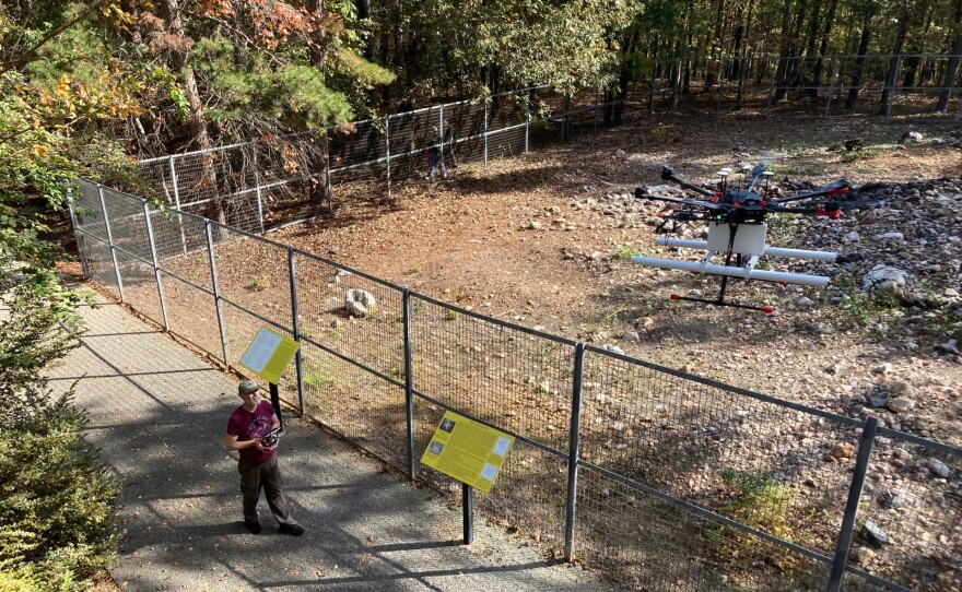 A person stands on a paved path holding a controller while a large multi-rotor drone hovers above.