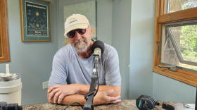 A bearded man in a blue shirt, sunglasses and baseball cap looks into the camera. He is sitting behind a microphone in a radio studio.