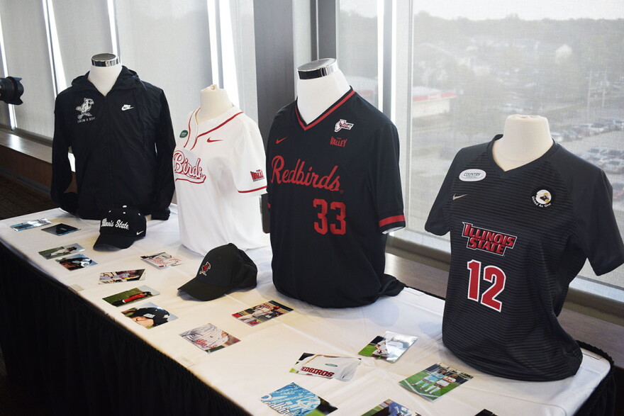 Four sports jerseys placed on mannikins on a table with photos and several caps. The uniforms are a mix of black, red and white and feature corporate logos on the shoulder. 