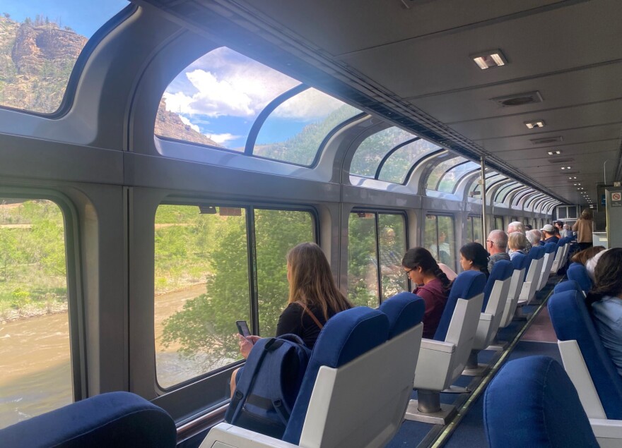 A row of passengers sits on a train, looking out the window. The train car has large picture windows, and windows on the curve of the train, on the side of the roof. The seats face out toward the big windows, with beautiful mountains outside.