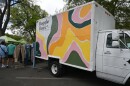 A classic box truck that’s painted with colorful swirls. On its left are two clothing racks with clothes and people walking alongside it. 