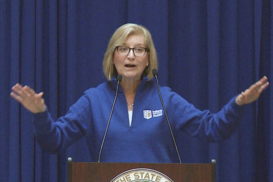 State Health Commissioner Dr. Kris Box stands in a blue Indiana Department of Health sweatshirt behind a podium with her arms out. She speaks about public health advancements in Indiana. 