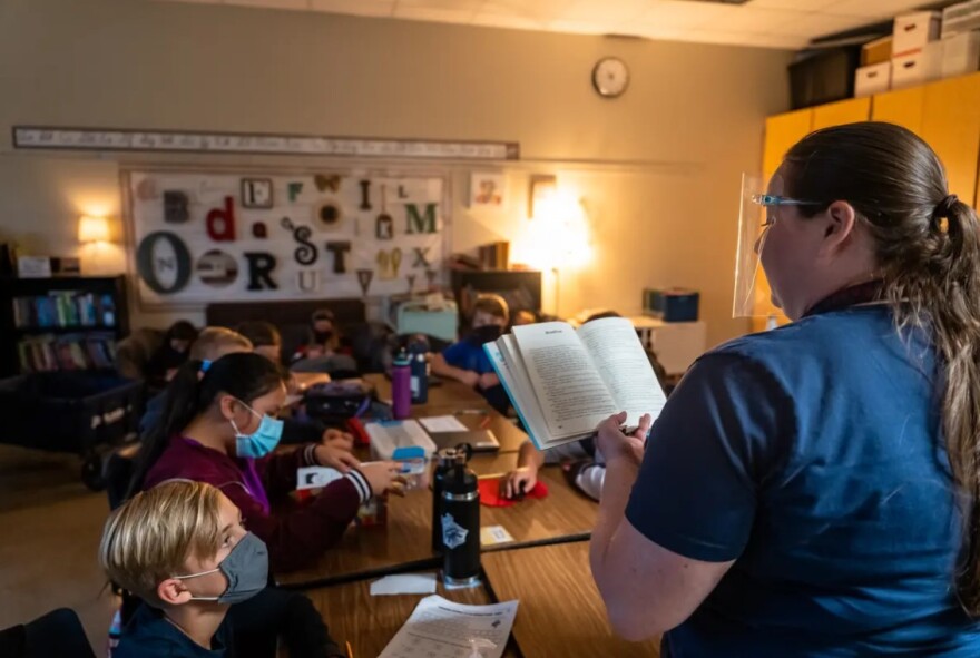 A teacher stands in front of her classroom, reading a book aloud to her students, who are wearing face masks.