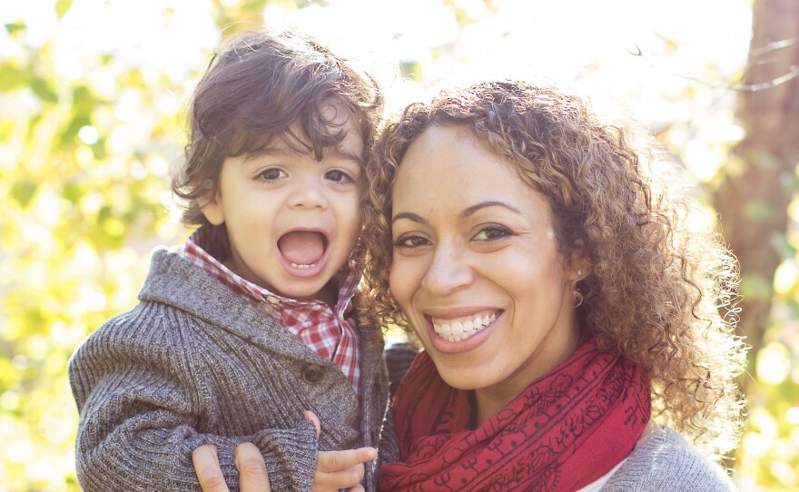 An african-American woman in a  white blouse and gray sweater with a red scarf stands outside in the sunshine with trees in the background and holds her son, a small boy of about 2 years. Both are smiling.