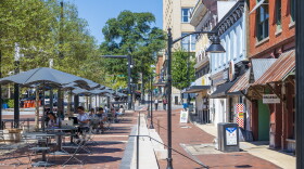 a view down a brick-paved pedestrian space on Aollege Square in Downtown Athens, GA. To the left is a row of umbrella-covered tables. To the right is a row of storefronts. Between them are small flights of three steps down from the tables to the storefronts.