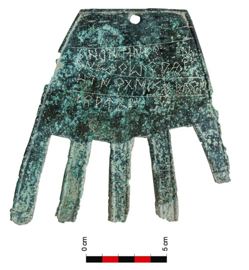 Set on a white background, the bronze hand is laid out with fingers facing down. It has a blue-greenish tint from oxidation. The words etched into it in dots are traced over the picture. A red and black scale next to it shows the hand is life-sized. 