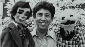 A black and white photo shows a smiling man flanked by two puppets, one puppet has a more human appearance while the other is furrier. The man is Ignacio Nacho Estrada.
