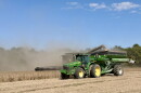 A green John Deere tractor pulling a wagon drives next to a combine harvesting soybeans.