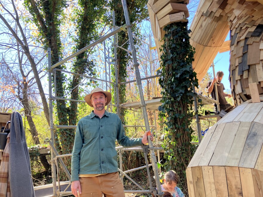 A wood sculptor stands outdoors next to his wooden statue.