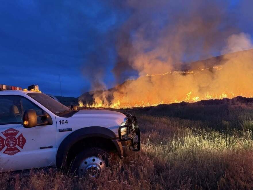 A white truck with a fire emblem on its side is parked at night in a field of tall grasses while a fire sends up flames a few feet tall a bit off in the distance.