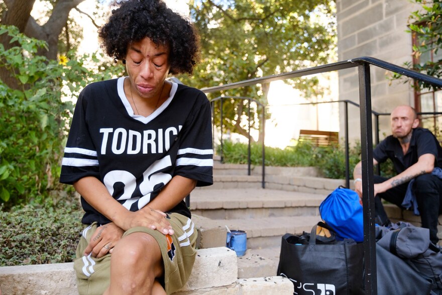 A person sits cross-legged outside on a concrete wall next to steps where another person sits with his belongings