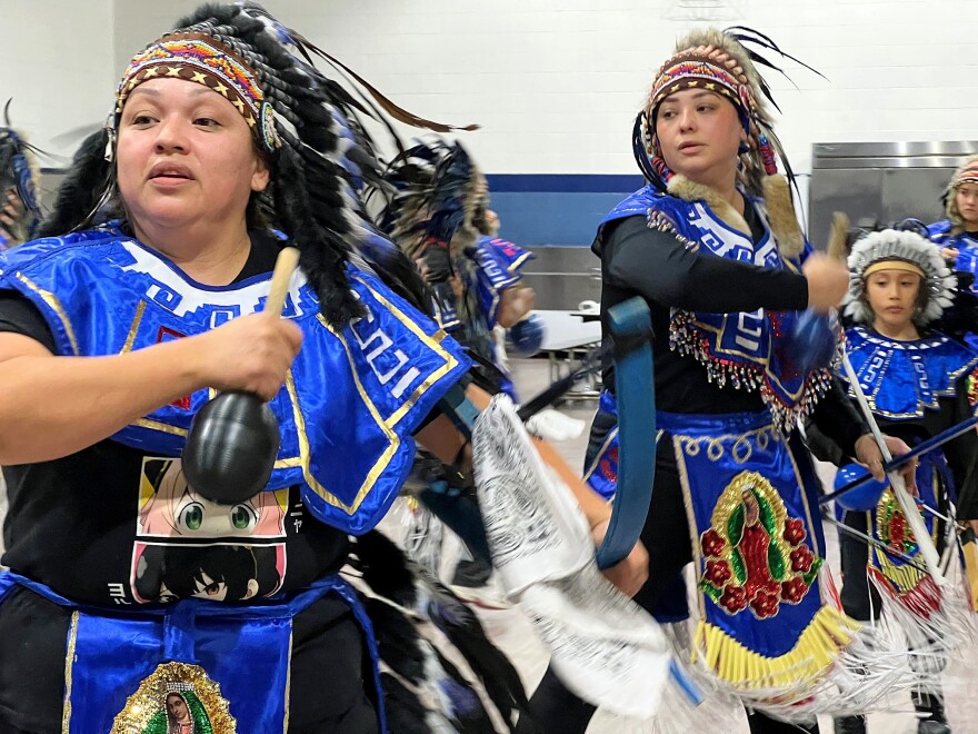 Dancers dressed in feathered headdresses and blue regalia spin and shake maraca-like instruments.