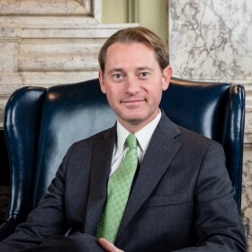 Michael Adams sitting in a leather chair, wearing a dark suit and green tie.