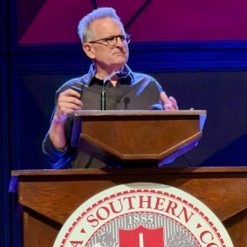 Man with gray hair and glasses standing behind a podium and speaking