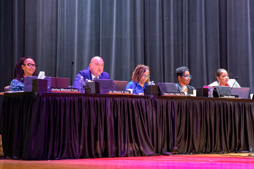 Five adults in suits sit in a line on an auditorium stage at a table with a black curtain in the background.