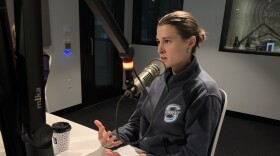 A woman in a gray fleece with the Seattle Torrent logo on it sits at a mic in a recording studio, talking and gesturing with her hands.