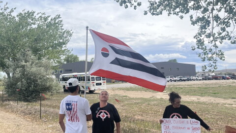 A boy in an American flag t-shirt holds up an Arapaho flag. He stands next to an older woman in a black t-shirt and another woman in a colorful skirt holding up a protest sign.
