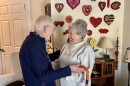 Elderly man stands with his cane and embraces his elderly wife, who smiles at him. They stand in a living room. A wall lined with pink and red wooden hearts is behind them.
