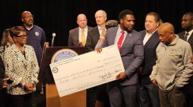 5/14 Memorial Foundation Executive Director Chad Houston, holding check, reacts to the foundation receiving $12 million in state funding. The money was presented by Assembly Majority Leader Crystal Peoples-Stokes, far-left, and Assembly Speaker Carl Heastie, far-right in gray jacket.