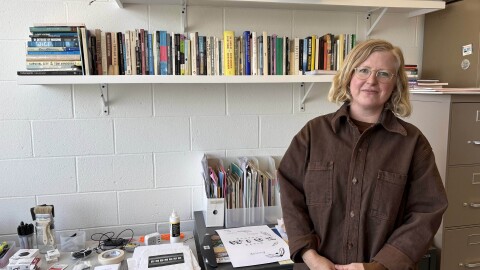 Carrie Hott in brown jacket standing in front of desk with papers and many tools and material. There is a bookshelf filled with books on the wall behind her.