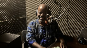 A man wearing headphones smiles while seated at a microphone.