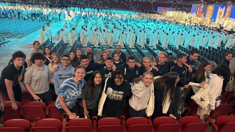 Several dozen high school students pose for a photo in an indoor arena as a gathering of men in white robes walk in a procession at a Catholic church gathering.